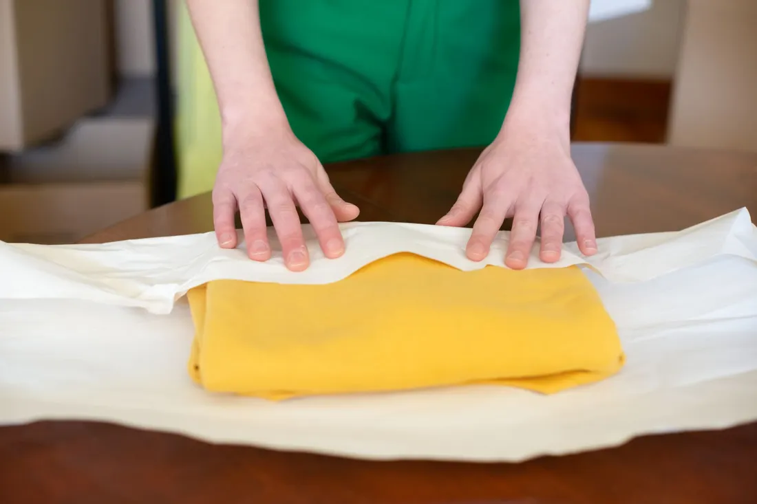 Hände glätten Packpapier über einem Stapel gefalteter Wäsche auf einem Tisch