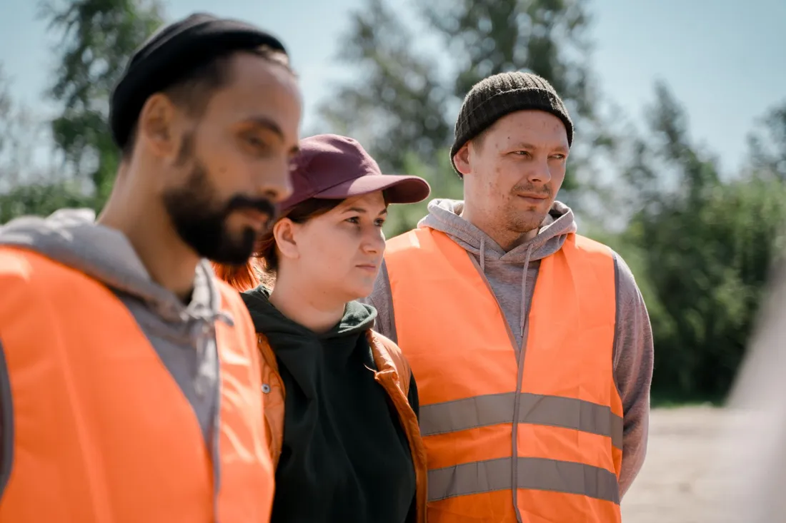 Drei Arbeiter in orangefarbenen Warnwesten stehen nebeneinander im Freien und blicken zur Seite