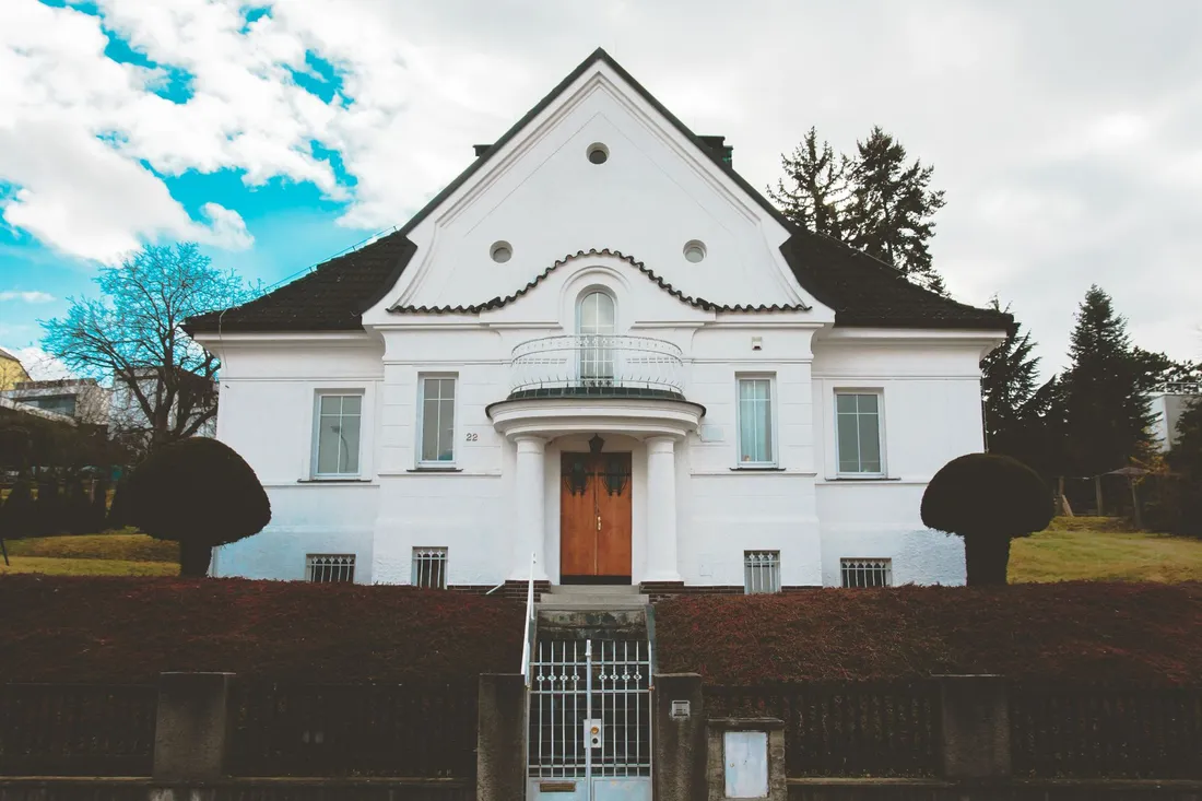 Weißes Einzelhaus mit Giebel und Rundbalkon hinter einer hohen Hecke und einem schmiedeeisernen Tor