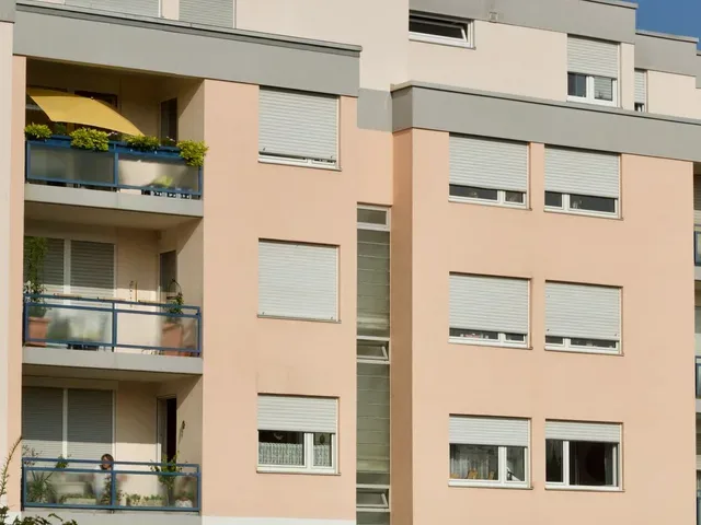 Mehrfamilienhaus mit Balkonen, Rollladenkästen und hellem Putz, davor Sträucher und Rasen