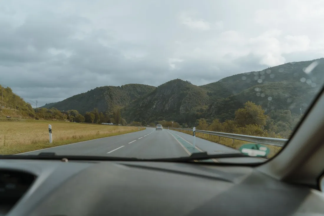 Blick durch die Windschutzscheibe auf eine Landstraße, die zwischen Wiesen auf bewaldete Berge zuläuft